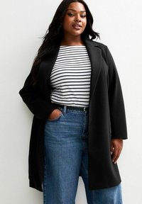 Zwarte lange jas met een gestructureerd ontwerp, gedragen over een wit met zwart gestreept shirt, gecombineerd met blauwe high-waisted jeans. Gladde stoftextuur.