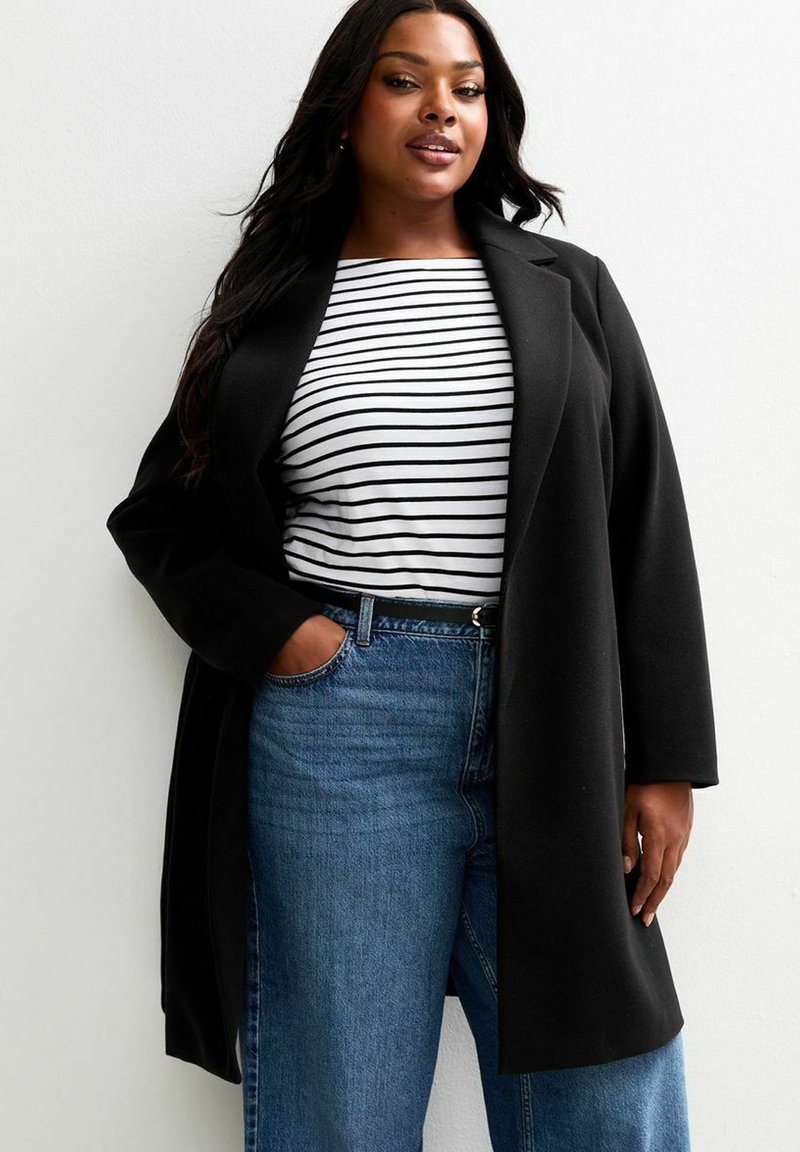 Zwarte lange jas met een gestructureerd ontwerp, gedragen over een wit met zwart gestreept shirt, gecombineerd met blauwe high-waisted jeans. Gladde stoftextuur.