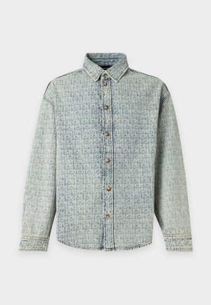 Noormees seisab käed taskutes, seljas heleroheline mustriline denim-särk ja sobivad laiad püksid valgete tossudega.