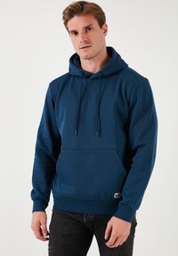 Dunkelblauer Hoodie aus weichem Material, mit einer frontalen Kängurutasche, Kordelzugkapuze und gerippten Bündchen. Schwarzes Etikett am Saum.