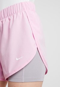 Vaaleanpunaiset kerrokselliset urheilushortsit, joissa on elastinen vyötärö ja valkoinen Nike-logo ulkokerroksessa harmaiden istuvien shortsien päällä.