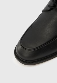 Μαύρο δερμάτινο παπούτσι με στρογγυλεμένη μύτη, ραφές κατά μήκος της άκρης και ομαλή υφή. Κορδόνια φαίνοντ αι στην κορυφή.