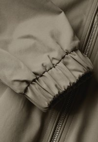 Hnědá látková manžeta bundy se sešitým elastickým detailem a kovovým zipem. Textura je hladká s mírným leskem.