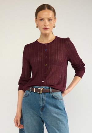 Femme portant un pull en tricot rouge foncé boutonné avec des boutons multicolores, un jean bleu, une ceinture marron et des boucles d’oreilles créoles dorées.