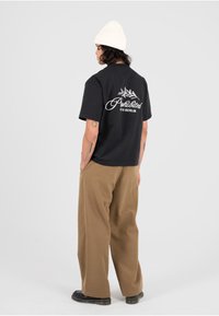 Czarny bawełniany t-shirt z białym graficznym napisem na plecach, zestawiony z brązowymi szerokimi spodniami i czarnym obuwiem, uzupełniony kremową dzianinową czapką.