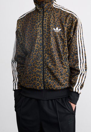 Braune und schwarze Leopardenmuster Adidas Trainingsjacke mit weißen Streifen an den Ärmeln, einem Frontreißverschluss und Adidas-Logo auf der Brust, getragen mit schwarzen Hosen.