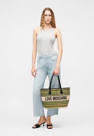 Femme aux longs cheveux ondulés portant un haut gris sans manches, un jean large bleu clair, des sandales noires à talons, tenant un sac cabas rayé Love Moschino.