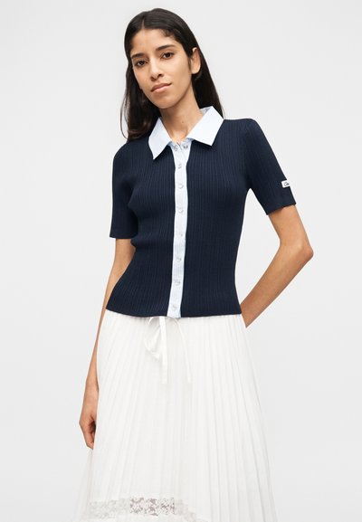 Γυναίκα που φοράει κοντομάνικο πλεκτό μπλε σκούρο τοπ με γαλάζιο γιακά και κουμπιά, συνδυασμένο με λευκή πλισέ φούστα με δαντέλα στο τελείωμα.
