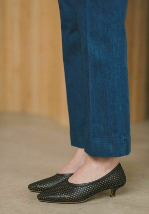 Pies con zapatos negros perforados de tacón bajo combinados con pantalones azules cortos sobre un fondo neutro.