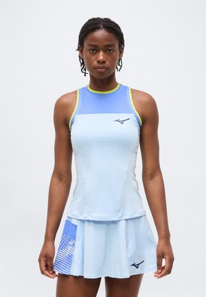 Γυναίκα που φοράει ανοιχτό μπλε αμάνικο αθλητικό τοπ και ταιριαστή πλισέ φούστα με σκούρο μπλε λογότυπο, στέκεται μπροστά σε απλό φόντο.