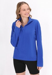 Pullover in pile blu con colletto alto, zip parziale sul davanti e maniche lunghe. Texture morbida; indossato con pantaloni corti neri.