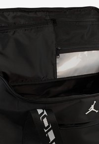 Μαύρη νάιλον τσάντα duffle με μεγάλο άνοιγμα, τσέπη από δίχτυ και λευκό λογότυπο Jumpman. Το εσωτερικό είναι λείο με τσέπη με φερμουάρ.