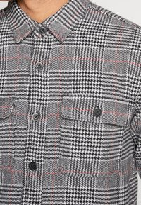 Närbild av en grå och svart houndstooth-mönstrad skjorta med röda linjer, knappar framtill och en bröstficka med knappflik.