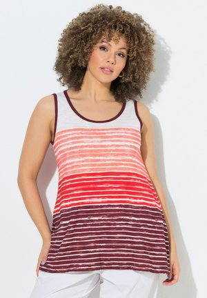 Frau mit lockigem Haar, die ein ärmelloses gestreiftes Top in den Farbtönen Weiß, Orange, Rot und Bordeaux zusammen mit weißen Hosen trägt und vor einer weißen Wand steht.