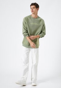 Sudadera verde con cuello redondo y puños acanalados, con texto en la parte delantera. Combinada con pantalones cargo blancos y zapatillas blancas.