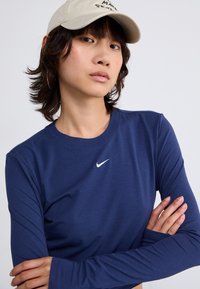 Tamsiai mėlynas ilgomis rankovėmis Nike marškinėlis su apvalia iškirpte ir balta logotipu. Lygi, elastinga medžiaga su ilgomis rankovėmis ir pritvirtintais rankogaliais.