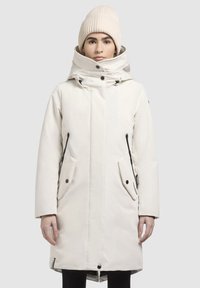 Witte geïsoleerde parka met hoge kraag, knoop- en ritssluitingen, zwarte accenten en grote zijzakken. Model draagt een gebreide beige beanie.