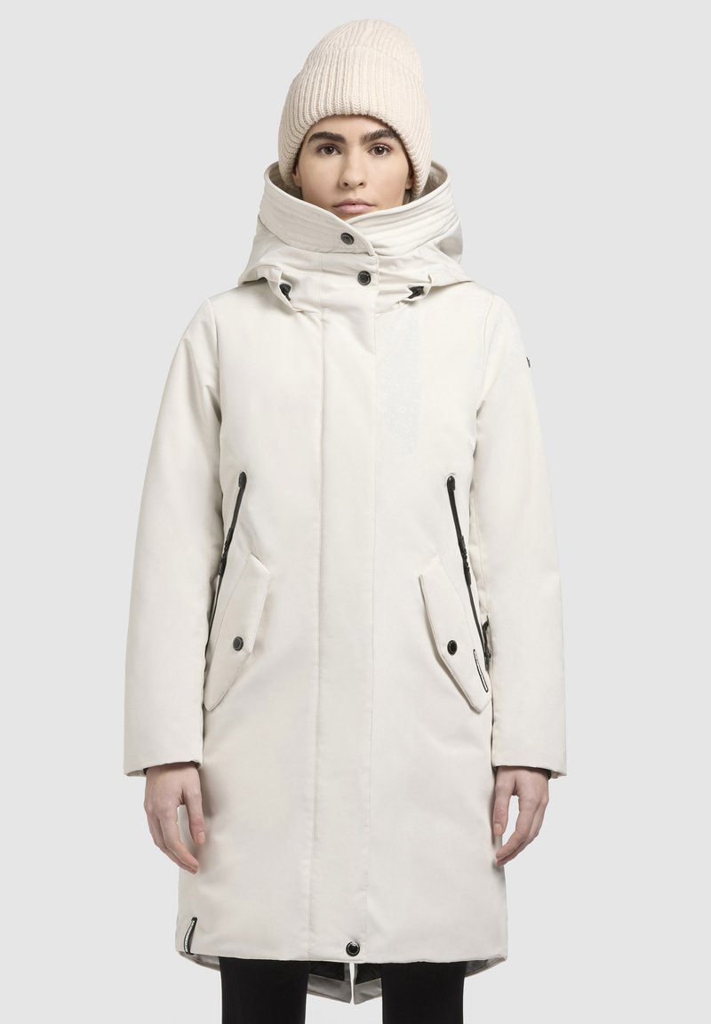 Witte geïsoleerde parka met hoge kraag, knoop- en ritssluitingen, zwarte accenten en grote zijzakken. Model draagt een gebreide beige beanie.