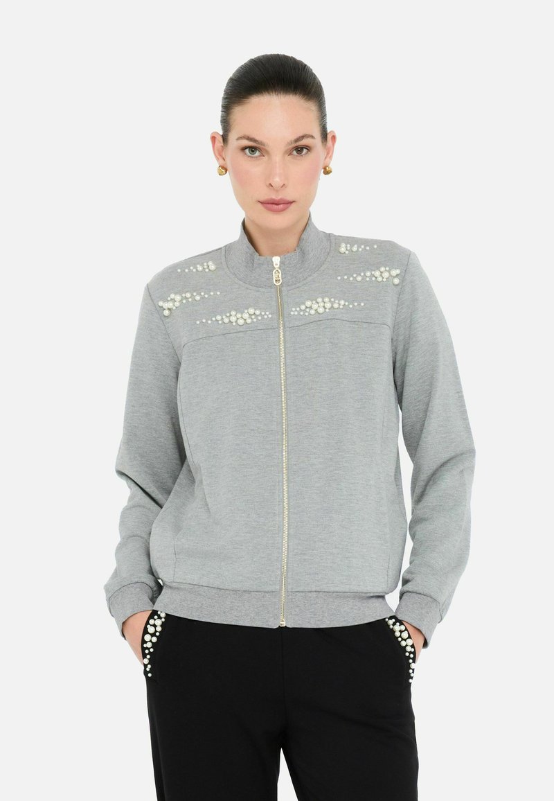 Felpa grigia con zip decorata con perle sul petto e colletto alto; abbinata a pantaloni neri con dettagli in perla sulle tasche.
