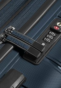 Gros plan d'une valise bleue texturée avec deux tirettes de fermeture éclair étiquetées Redolz et une serrure TSA à combinaison à trois chiffres intégrée.