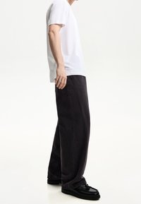 Mann i en enkel vit t-shirt, lösa svarta jeans och svarta läderloafers som står mot en vit bakgrund.