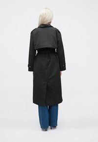 Zwarte trenchcoat met een kort ontwerp, ceintuur in de taille en decoratieve knopen op de schouders. Draag het over blauwe wijde jeans.