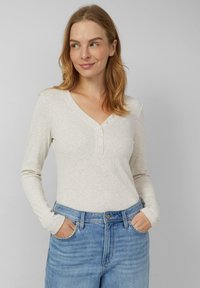 Jasnobeżowa koszulka henley z długim rękawem i dekoltem w serek, zapinana na trzy przednie guziki. Połączona z niebieskimi, wysokim stanem jeansami.