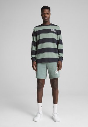 Chemise à manches longues rayée verte et marine avec un logo, associée à un short vert menthe avec impression de logo, portée avec des baskets blanches.