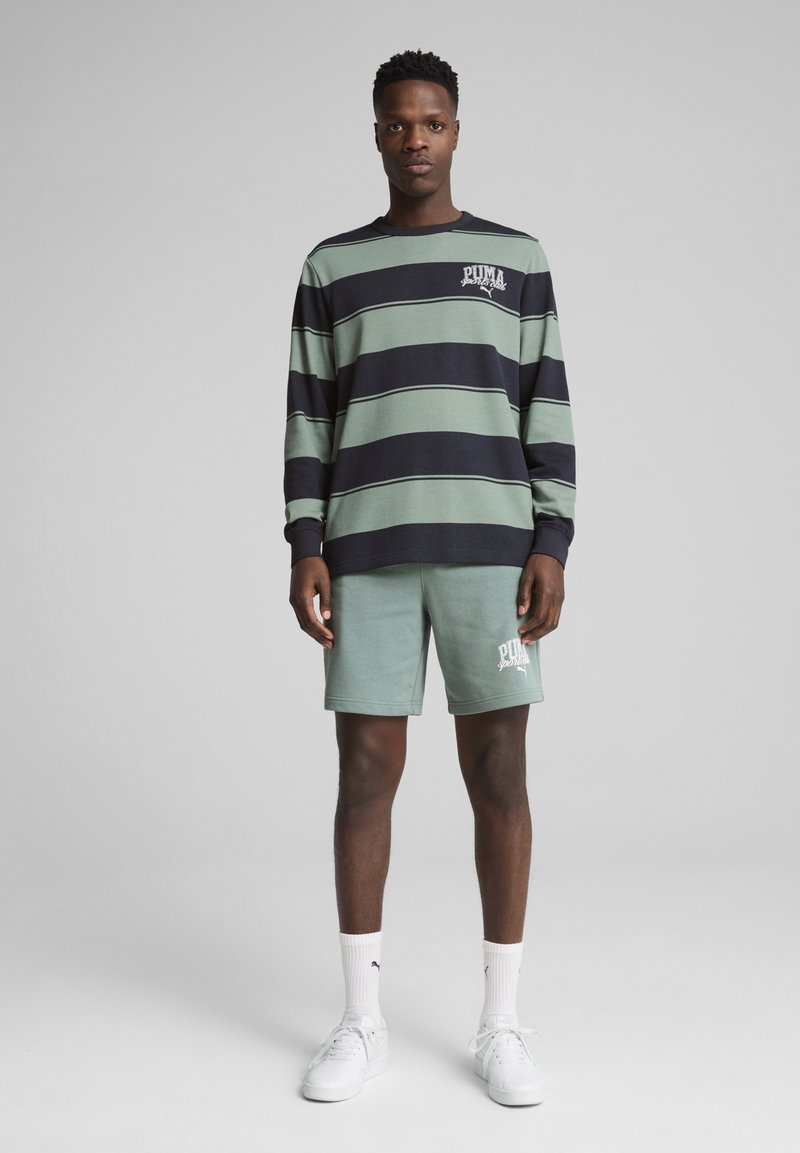 Langsleeve gestreepte shirt in groen en marineblauw met een logo, gecombineerd met mintgroene shorts met logo-print, gedragen met witte sneakers.