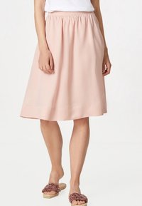 Nainen, jolla on päällään vaaleanpunainen, polvipituinen hame, jossa on joustava vyötärö, valkoinen yläosa ja pörröiset vaaleanpunaiset sandaalit.