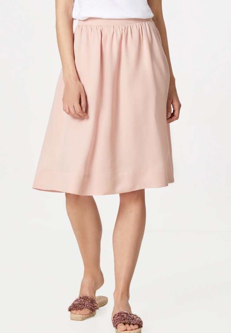 Kvinna klädd i en ljusrosa, knälång kjol med elastisk midja, vit topp och fluffiga rosa sandaler utan klack.