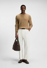 Beige Kabelstrickpullover, weiße weit geschnittene Hose, braune Lederschuhe und eine dunkelbraune Handtasche mit silbernen Beschlägen, vor einem neutralen Hintergrund.
