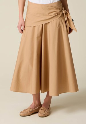 Jupe midi beige en ligne A avec détail de nœud sur le côté portée avec des mocassins beige assortis sur une personne debout.