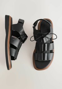 Mustat nahkasaappaat leveillä hihnoilla, teksturoidulla pinnalla ja ruskealla kumipohjalla; mukana nauhoitusyksityiskohta ja säädettävä takahihna.