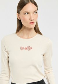 Lange mouw, crèmekleurig top met een rood bloemenvisontwerp op de borst. Gemaakt van ribstoffen, aansluitende pasvorm.