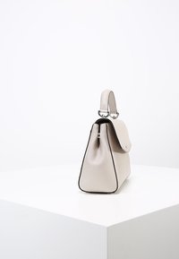 Widok z boku beżowej skórzanej torebki o sztywnej formie, z kontrastującym czarnym wykończeniem oraz krótkim uchwytem z metalowymi elementami.