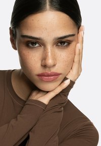 Brun långärmad topp med figursydd design, subtil glans och textilstruktur. Modellen har mörkt hår och fräknar, med handen vila mot kinden.