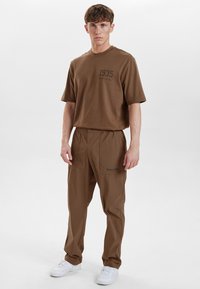 Brun bomulds t-shirt med "1935" tryk, parret med brune bukser med lommer og logo broderi, kompletteret med hvide sneakers.