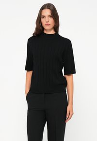 Svart kortermet strikket topp med ribbestruktur, høy hals og en tettsittende design. Kombinert med svarte bukser for et monokromt utseende.