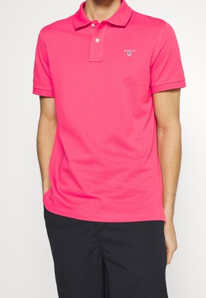 Homme portant un polo rose vif à manches courtes avec boutons et pantalon noir, debout avec une main dans la poche.