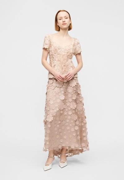 Γυναίκα που φοράει ροζ παλ τοπ με υφή και κοντά μανίκια με λουλουδένιο μοτίβο και μακριά φούστα με τρισδιάστατα λουλουδένια σχέδια, συνοδευόμενο από λευκά μυτερά παπούτσια.