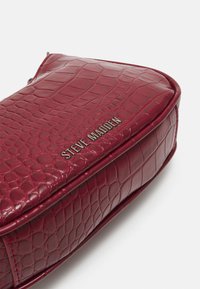Burgunderfarget faux skinnveske med et strukturert krokodillemønster, med en gullfarget "Steve Madden"-logo på siden, rund form.