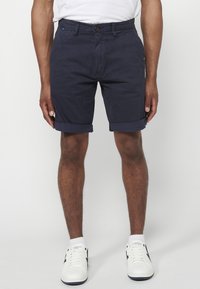 Marineblaue Baumwollshorts mit umgeschlagenem Saum, Fronttaschen und Knopfverschluss. Kombiniert mit weißen Sneakers mit dunklen Akzenten.