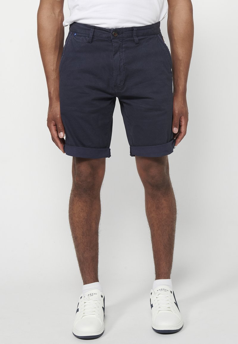 Marineblaue Baumwollshorts mit umgeschlagenem Saum, Fronttaschen und Knopfverschluss. Kombiniert mit weißen Sneakers mit dunklen Akzenten.