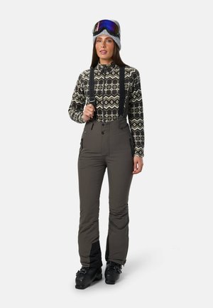 Femme portant un haut zippé à motif, pantalon de ski gris avec bretelles, chaussures de ski noires, bonnet gris et lunettes de ski bleues sur le front.