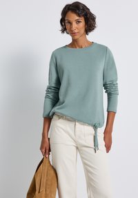 Femme aux cheveux courts et bouclés portant un haut à manches longues teal clair et un pantalon écru, tenant un sac en suède beige contre un fond uni.