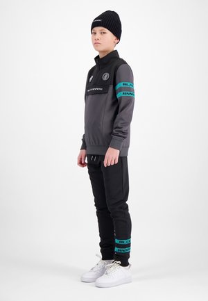 Jong persoon staand, gekleed in een zwarte muts, een grijs-zwarte sportjas met teal strepen, zwarte broek en witte sneakers tegen een witte achtergrond.