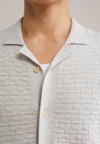 Cardigan grigio chiaro, testurizzato con motivi geometrici, dotato di un colletto morbido e un singolo bottone in madreperla. Materiale in maglia liscia.