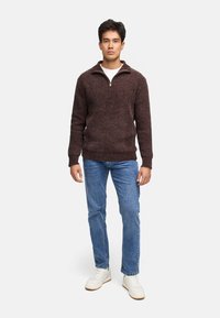 Brun zip-up sweater med et tekstureret strikmønster, parret med lyseblå jeans og hvide sneakers, som står foran en ensfarvet baggrund.