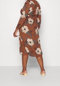 Kvinde iført en brun blomsteret midi kjole og beige blokhæle sandaler, som står foran en ensfarvet hvid baggrund.